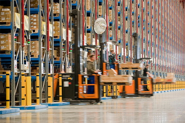 Gabelstapler fahren durch ein Hochregallager mit vollen Regalen und Kartons im laufenden Betrieb.