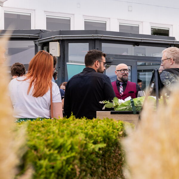 Mehrere Personen stehen an einer modernen Bar vor einer Bordwand mit Glasfronten, teilweise verdeckt durch grüne Sträucher im Vordergrund