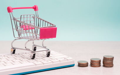 Mini-Einkaufswagen auf Tastatur neben gestapelten Münzen als Symbol für E-Commerce-Kosten